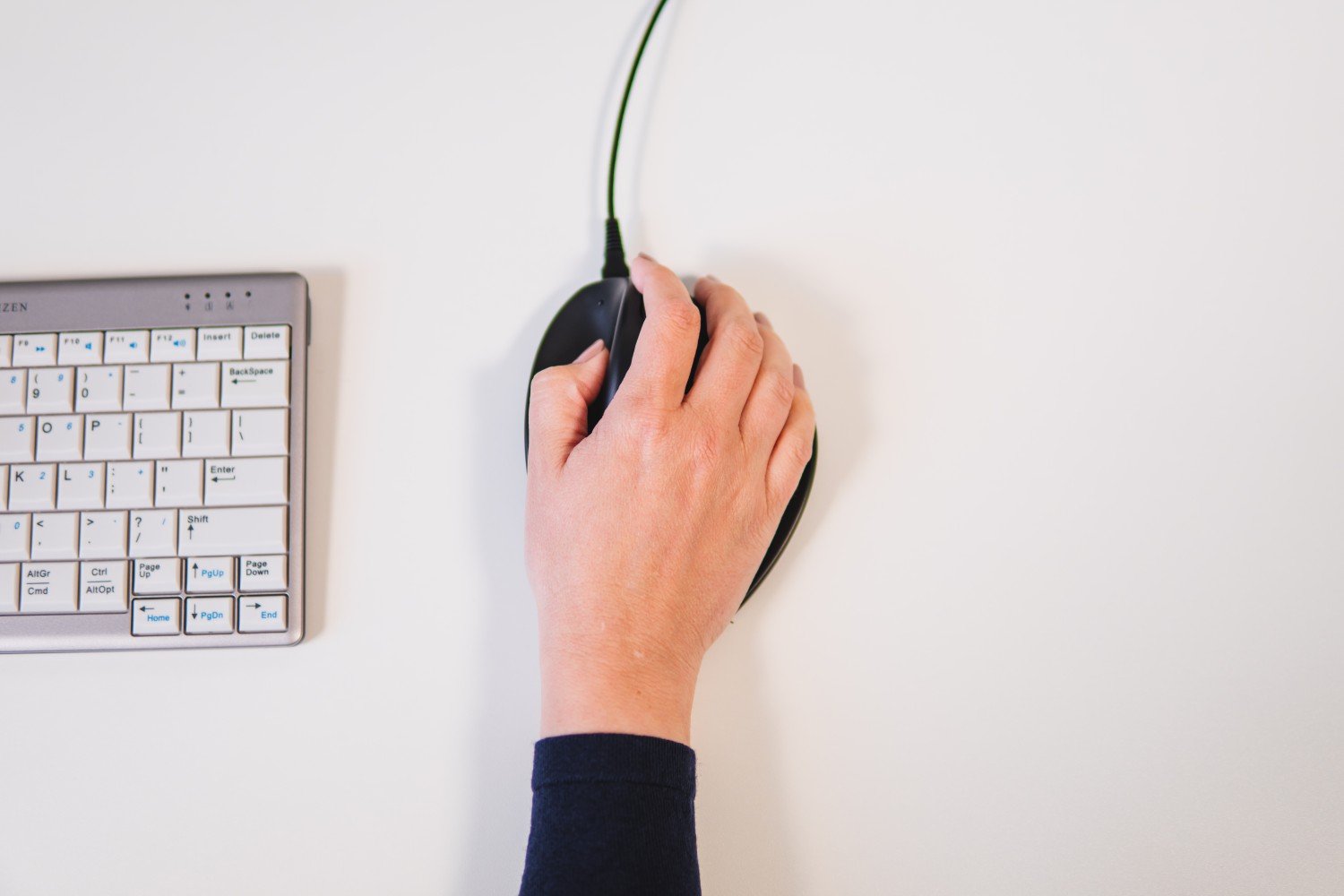 Hand met lichte huid die een zwarte computermuis vastpakt op een witte ondergrond naast een compact wit toetsenbord.