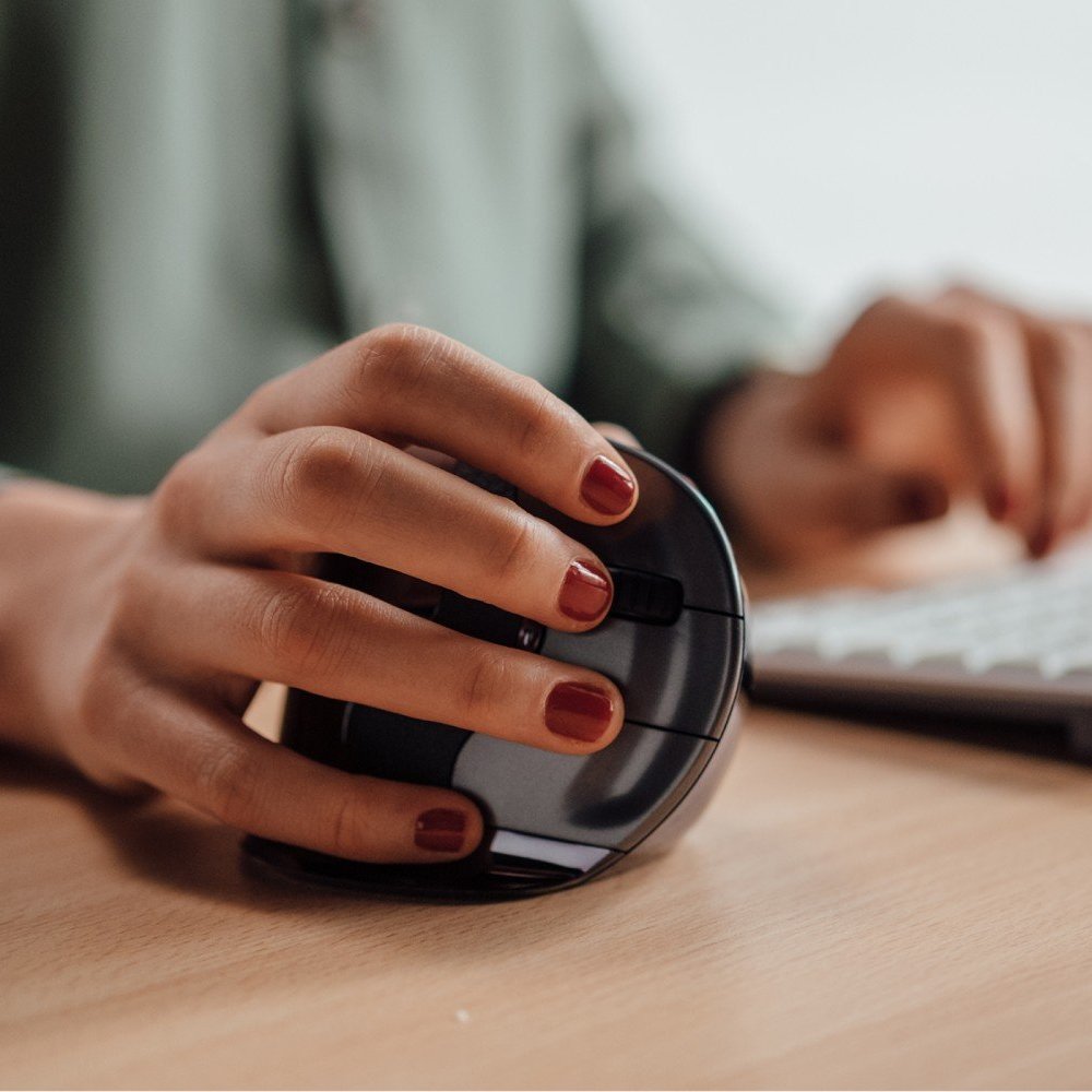 Hand met rood gelakte nagels gebruikt een verticale ergonomische computermuis op een houten bureau met een toetsenbord op de achtergrond.