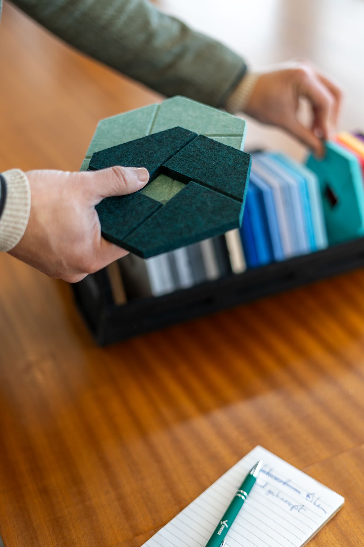 Handen houden meerdere monsters van akoestisch vilt in verschillende blauwtinten, met een zwart kistje met meer stalen en een notitieboekje met een groene pen op een houten tafel.