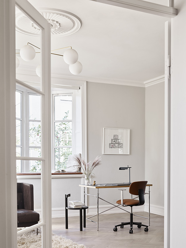Lichte thuiskantoorruimte gezien door een open deur, met een minimalistisch bureau, een oranje bureaustoel op wieltjes, een kleine zwarte bijzettafel met boeken en een vaas met bloemen, grote ramen met uitzicht op buiten, en een modern plafondlampje.