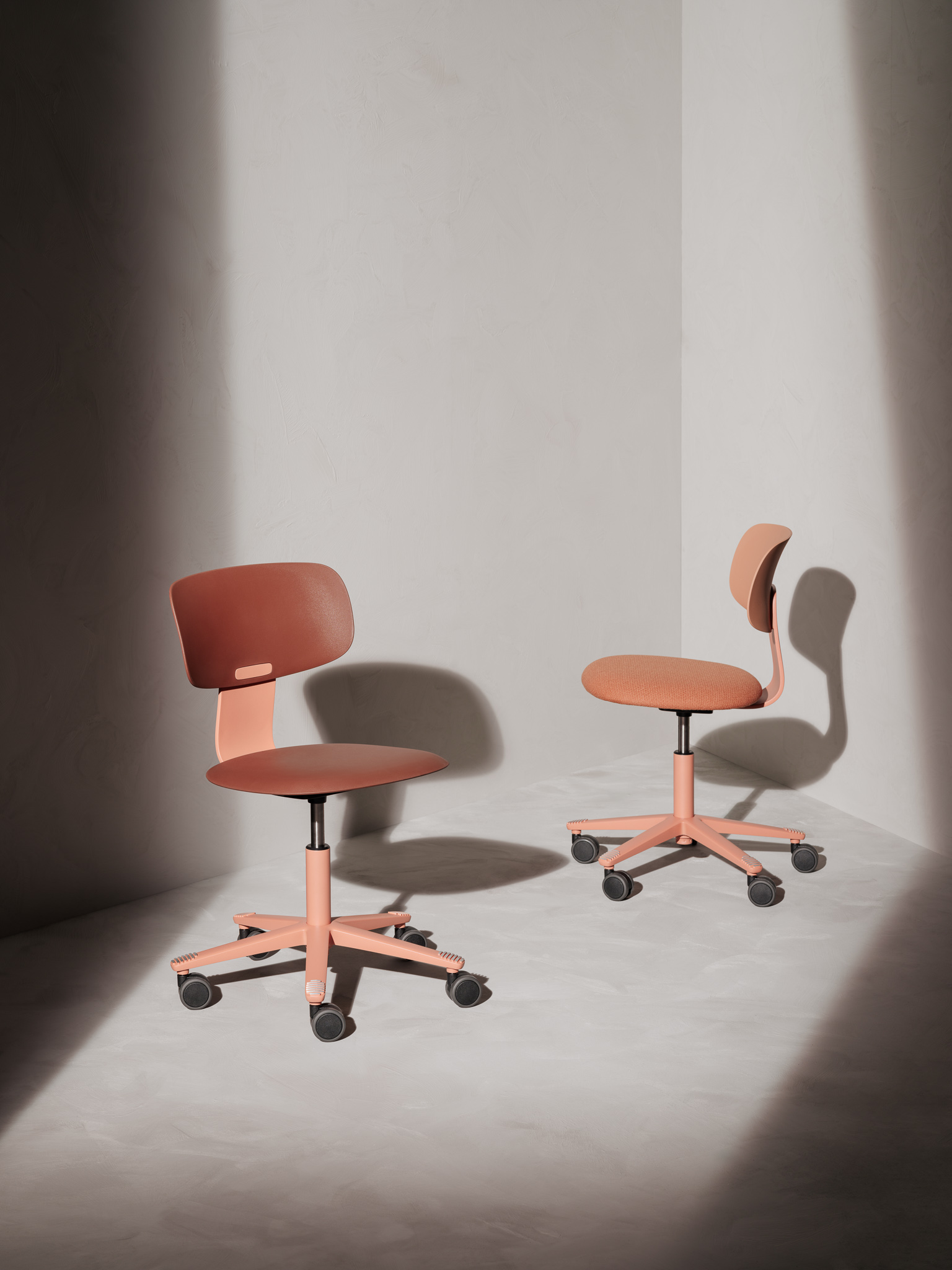 Twee oranje-bruine bureaustoelen met wielen staan in een hoek van een lichte kamer met schaduwen op de muur en vloer.
