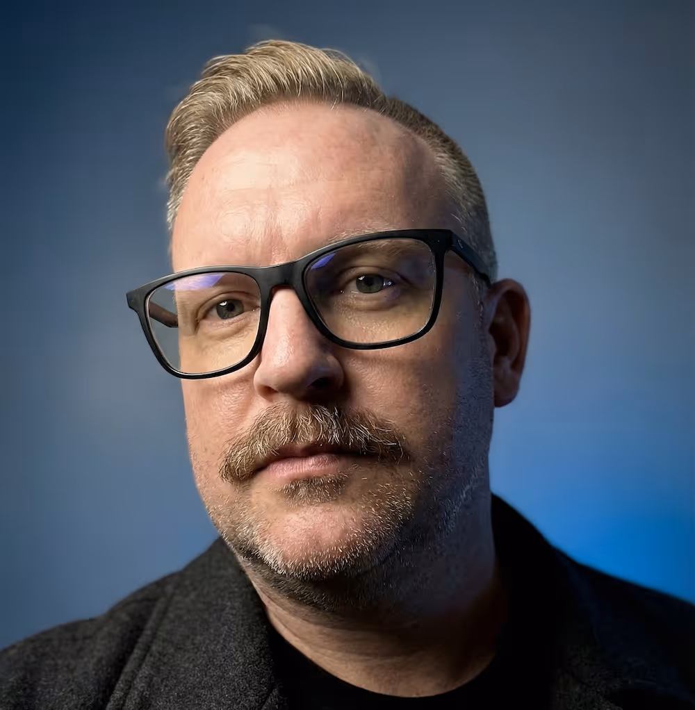 Close-up portrait of a middle-aged man with light hair, mustache, glasses, and a dark jacket against a blue background.