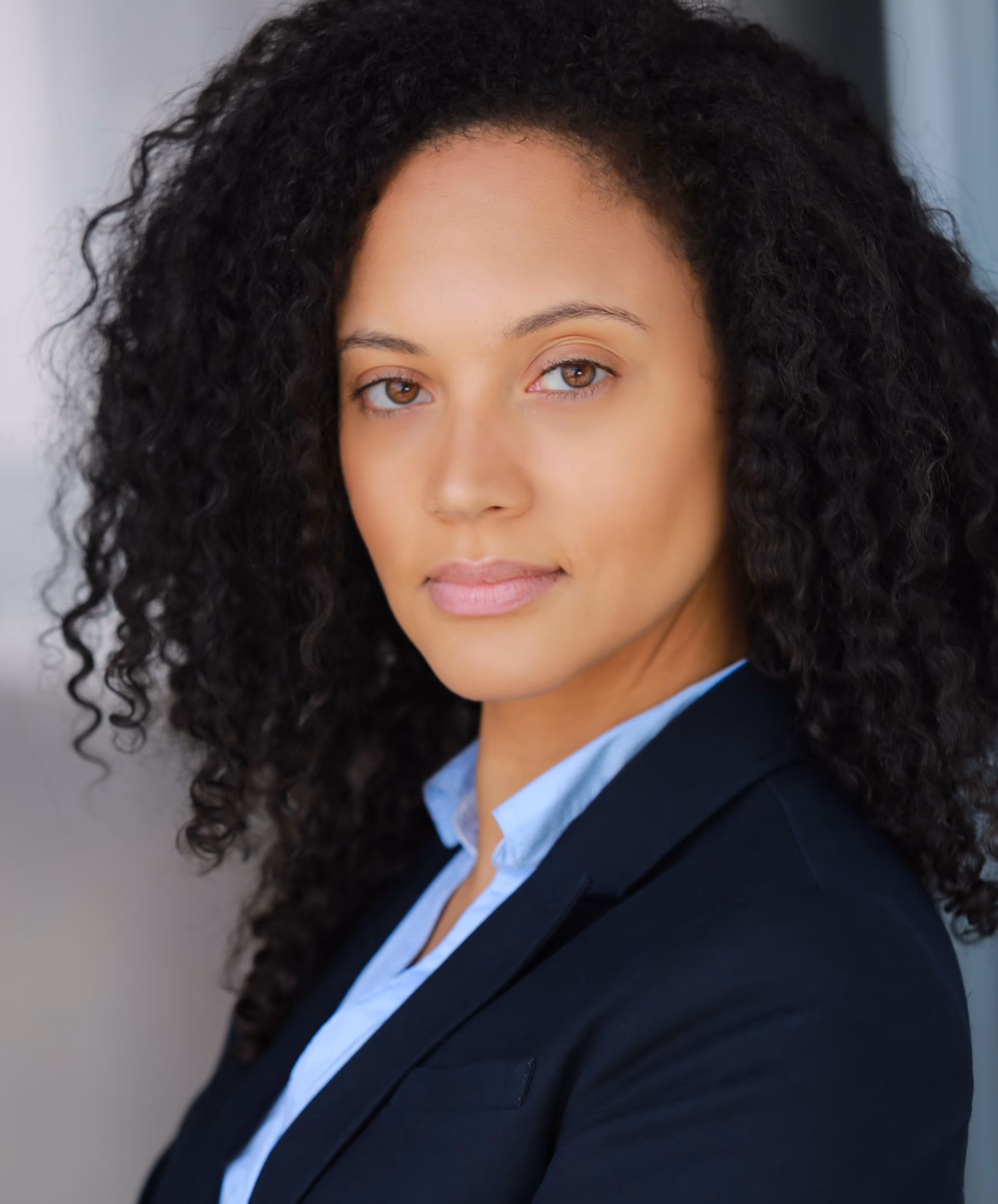 Professional woman with curly black hair wearing a navy blazer and light blue shirt looking confidently at the camera.