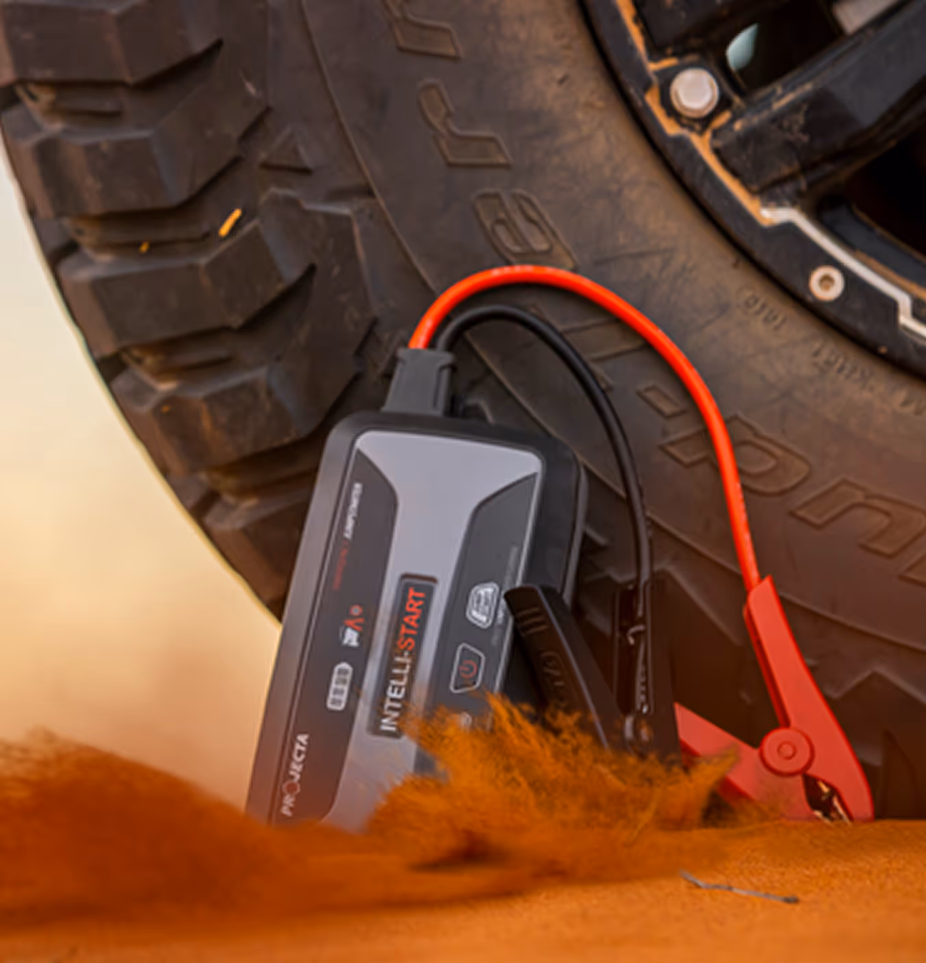 Portable jump starter with red and black clamps attached to a rugged vehicle tire on sandy ground.