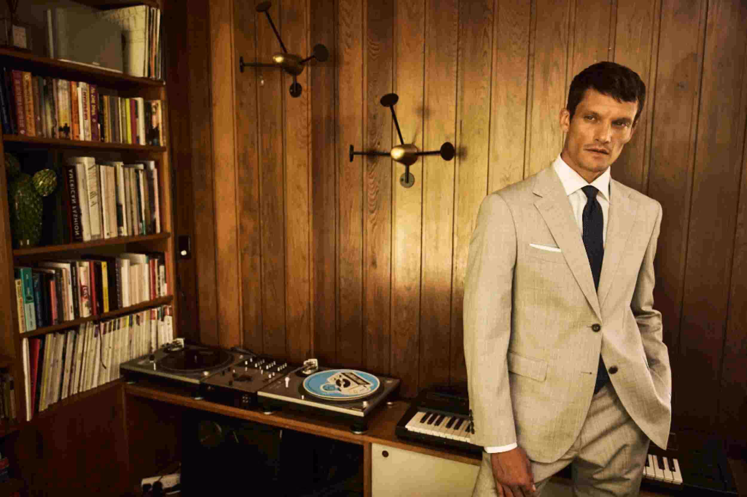 Man in a beige suit with a dark tie standing in a wood-paneled room with a turntable, keyboard, and bookshelf.