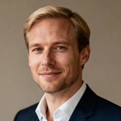 Portrait of a smiling blond man in a dark suit jacket and white shirt against a neutral background.