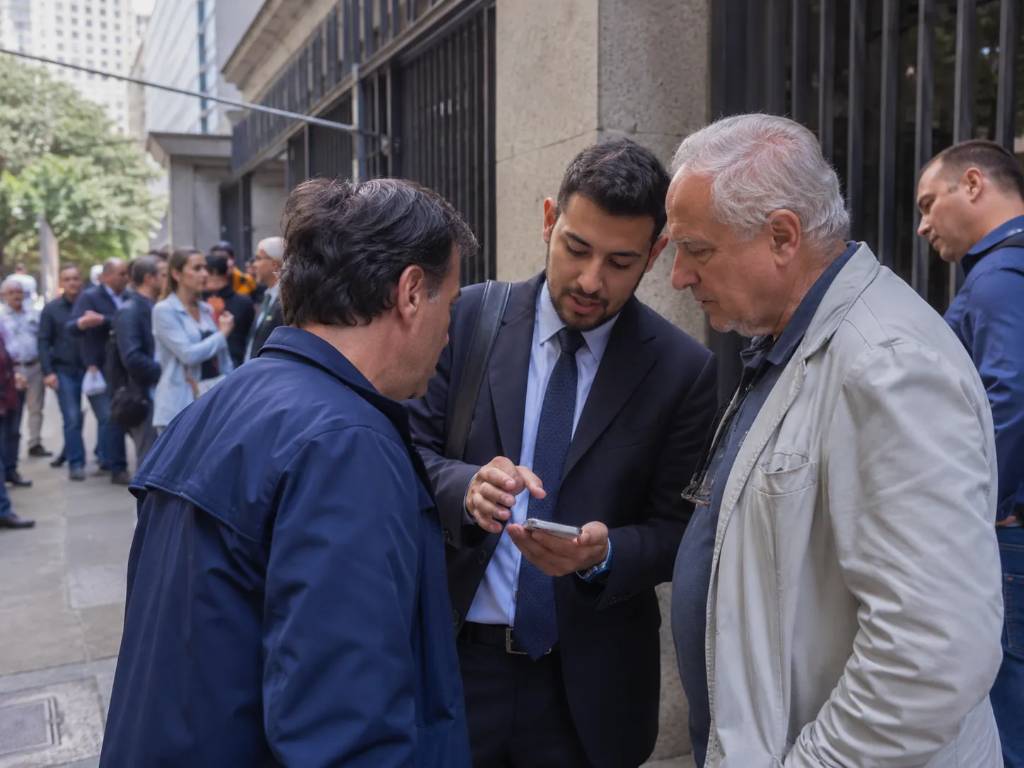 Tradutor prestando assistência a dois cidadãos italianos em frente a um consulado em São Paulo, em ambiente urbano movimentado.