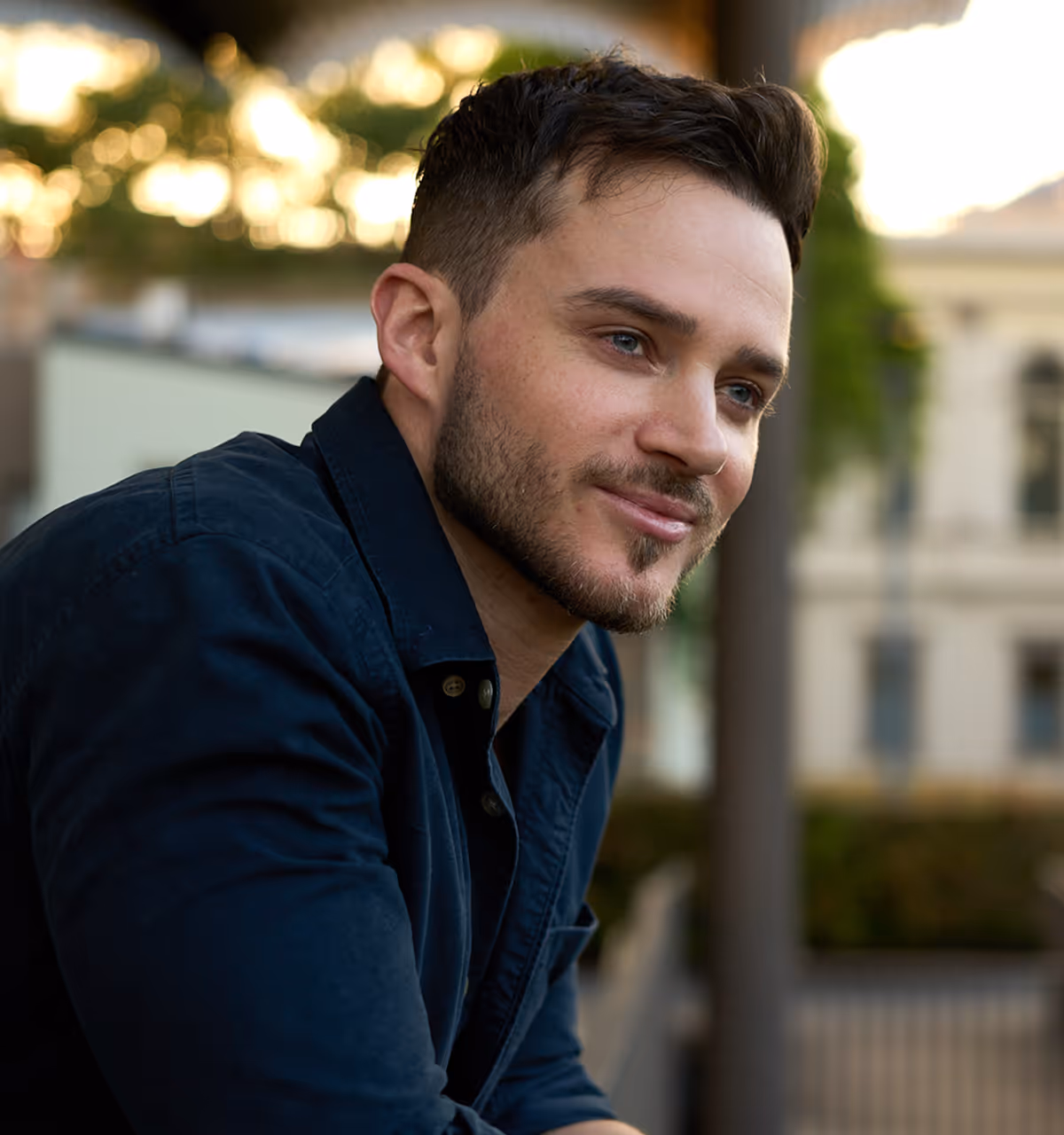 Portrait of a young man with short dark hair and a trimmed beard wearing a black button-up shirt against a dark textured background.