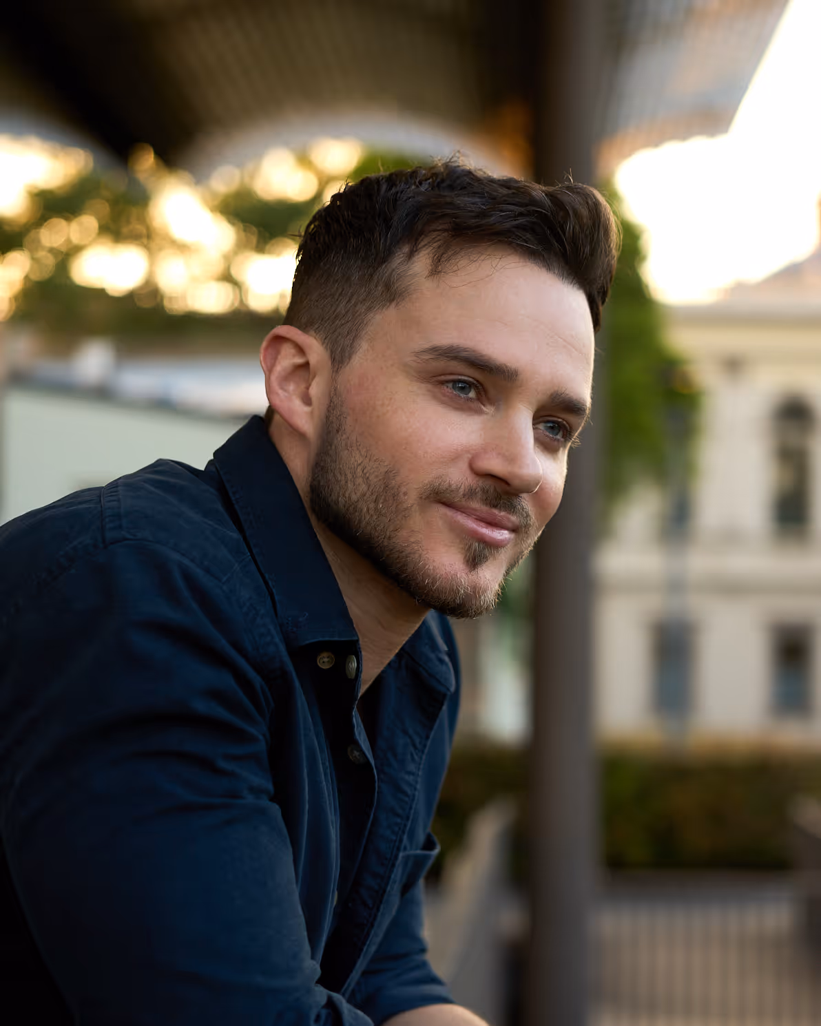 Portrait of a young man with short dark hair and a trimmed beard wearing a black button-up shirt against a dark textured background.
