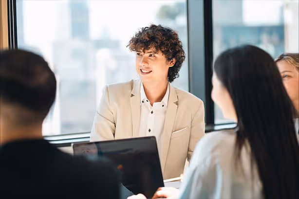 Young man with curly hair in a beige jacket smiling and sitting at a meeting table with colleagues in an office.