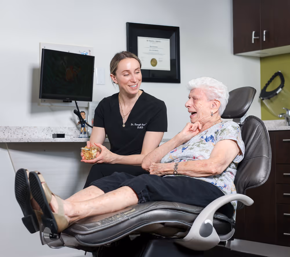 Dentist smiling and holding a model while an elderly woman laughs in a dental chair.
