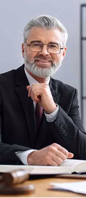 Smiling older man with gray hair and beard in glasses and suit sitting at desk with book and gavel.