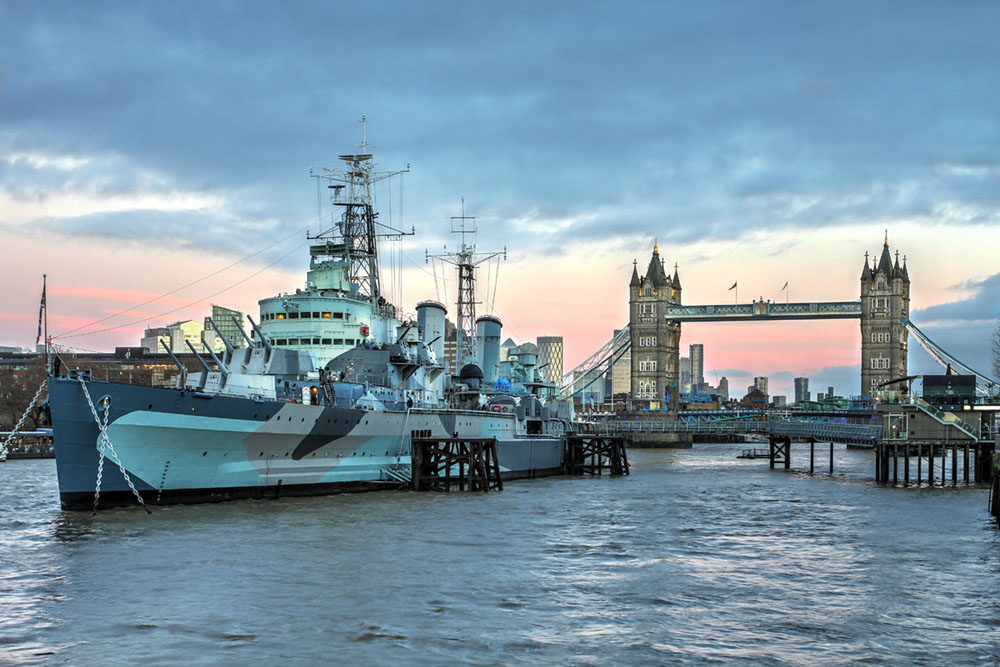 A wide shot of HMS Belfast, a camouflaged historic naval warship, moored on the River Thames. In the background to the right, London's iconic Tower Bridge stretches across the water under a cloudy, twilight sky with hints of pink and blue.
