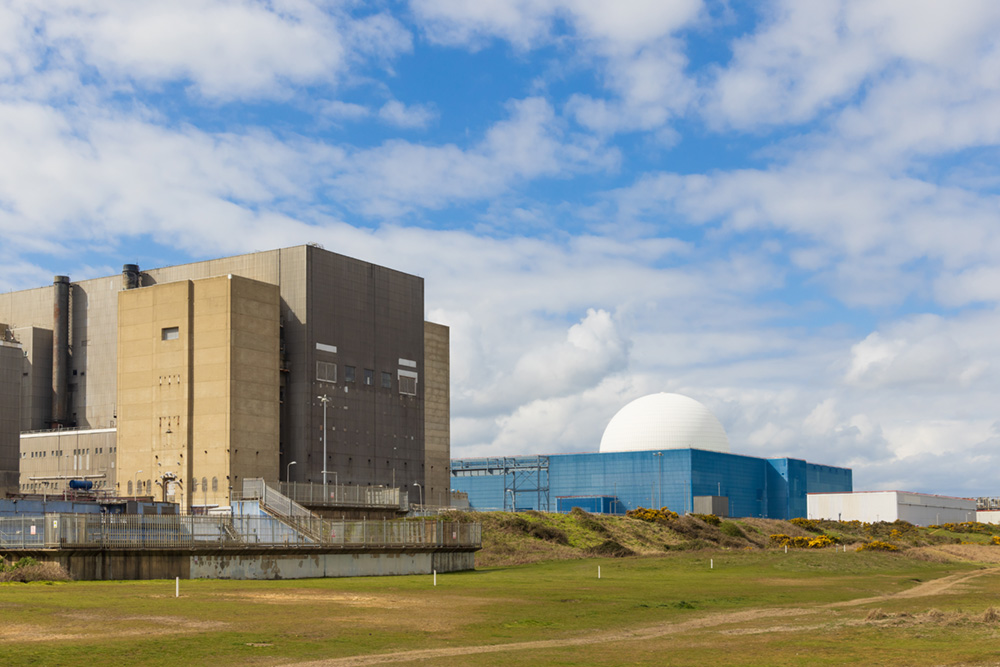 A nuclear power plant facility featuring a tall, blocky concrete building on the left and a blue building topped with a large white spherical dome on the right. The structures are set in a flat, grassy landscape under a bright blue sky with scattered white clouds.