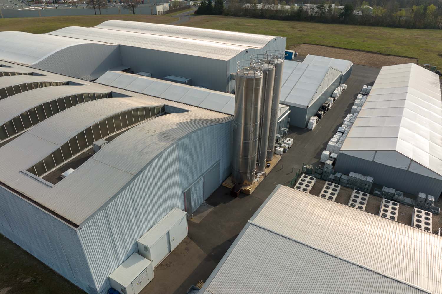 An aerial view of an industrial complex featuring large warehouse buildings with curved metal roofs, three tall silver storage silos, and neatly stacked pallets of materials in the outdoor staging area, surrounded by green fields.
