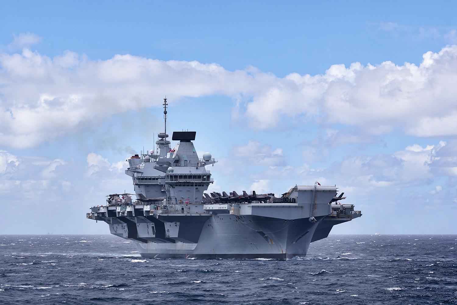 A large, gray Queen Elizabeth-class aircraft carrier sailing on the open sea under a blue sky with scattered white clouds. The ship features its distinctive dual-island design, a ski-jump ramp at the bow, and several aircraft parked on the flight deck.