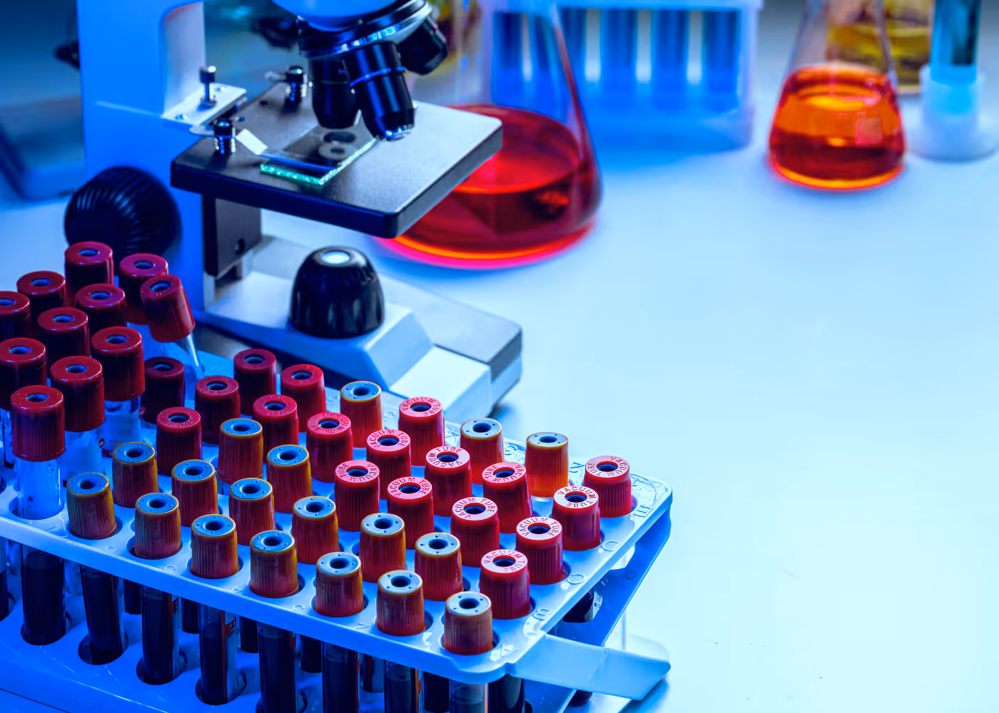 Laboratory microscope with test tubes and beakers containing colorful liquids on a lab bench.