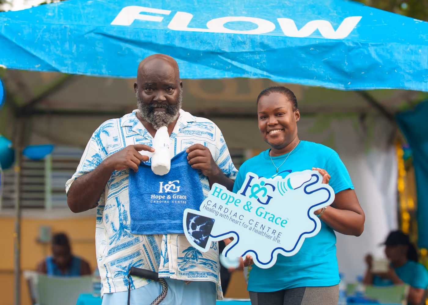 Two people holding Hope & Grace Cardiac Centre items under a tent with a blue 'FLOW' banner.