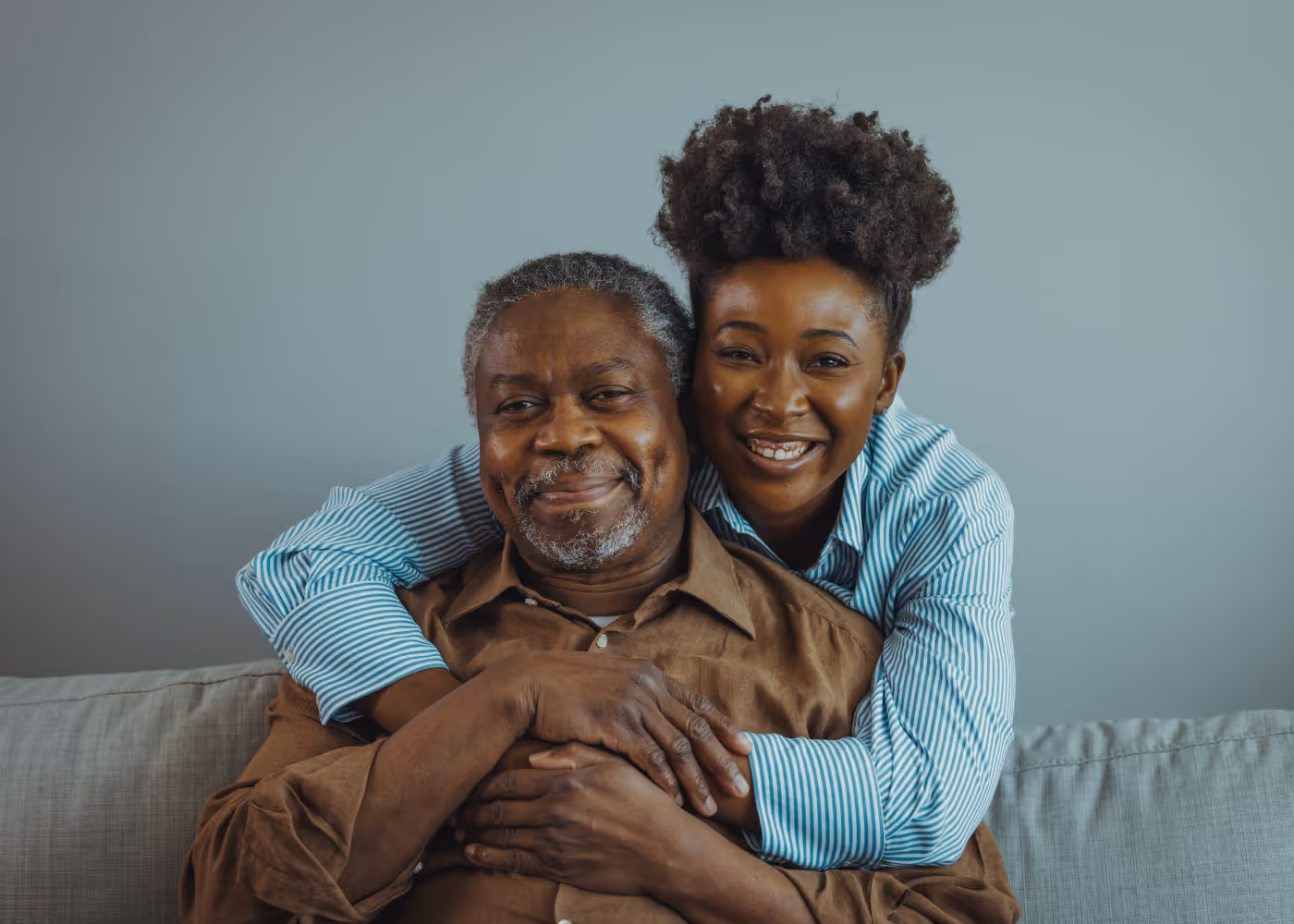 Smiling woman hugging elderly man from behind on a gray sofa with a plain gray wall behind them.