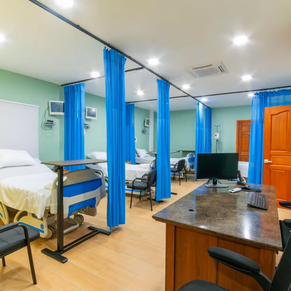 Hospital room with multiple beds separated by blue curtains and a desk with a computer.