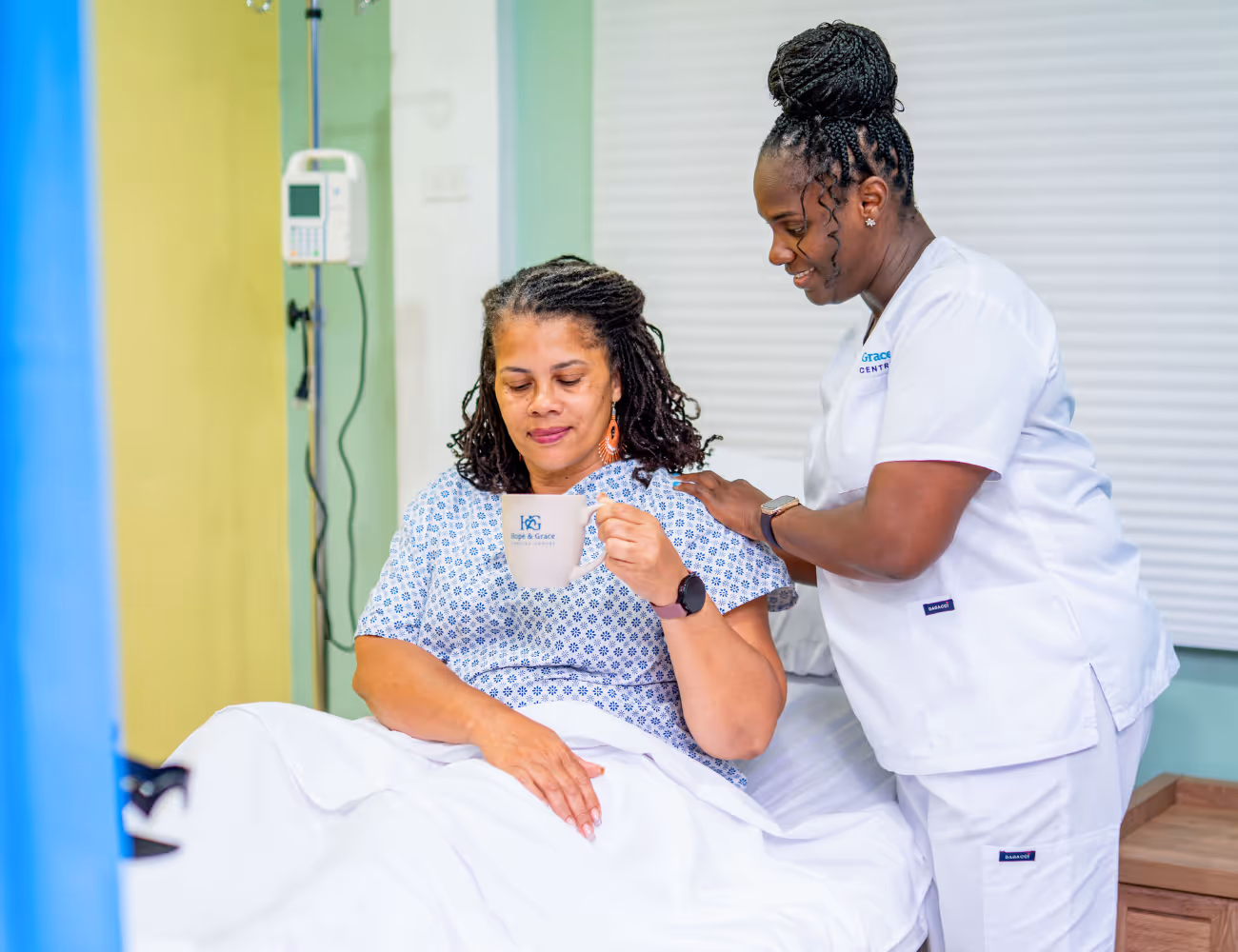 Nurse in white scrubs comforting seated patient holding a mug in a hospital room.