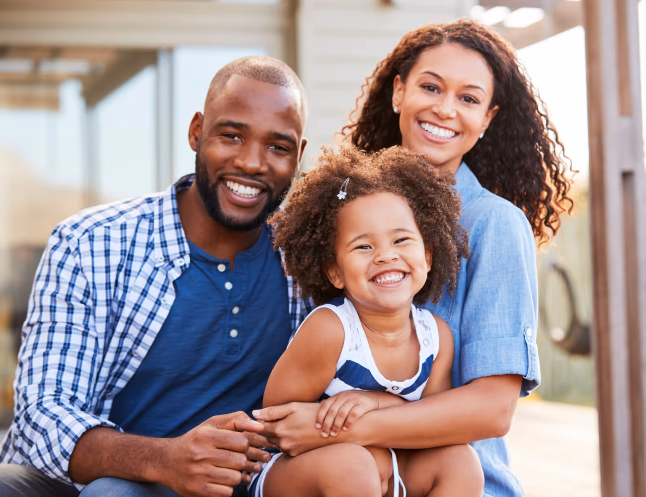 Smiling parents hug their happy young daughter outdoors in a bright setting.
