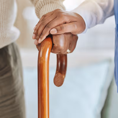 One hand gently holding another hand resting on a wooden cane, symbolizing care and support.