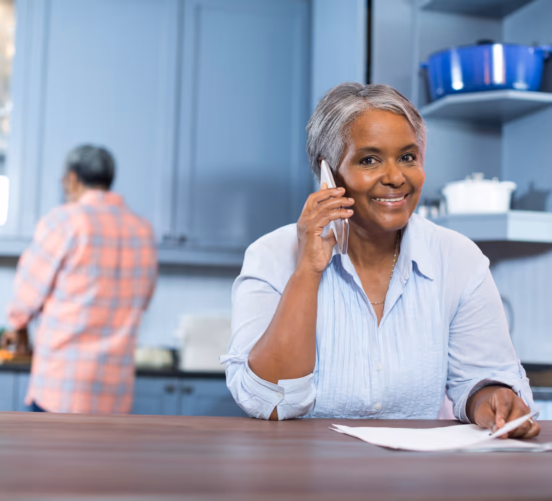 Smiling older woman talking on phone in kitchen while man in plaid shirt is blurred in background.
