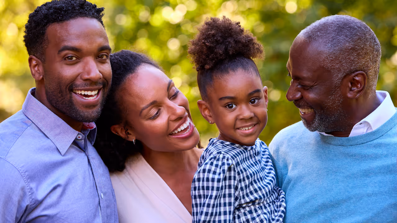 Four African American family members smiling outdoors with trees blurred in the background.