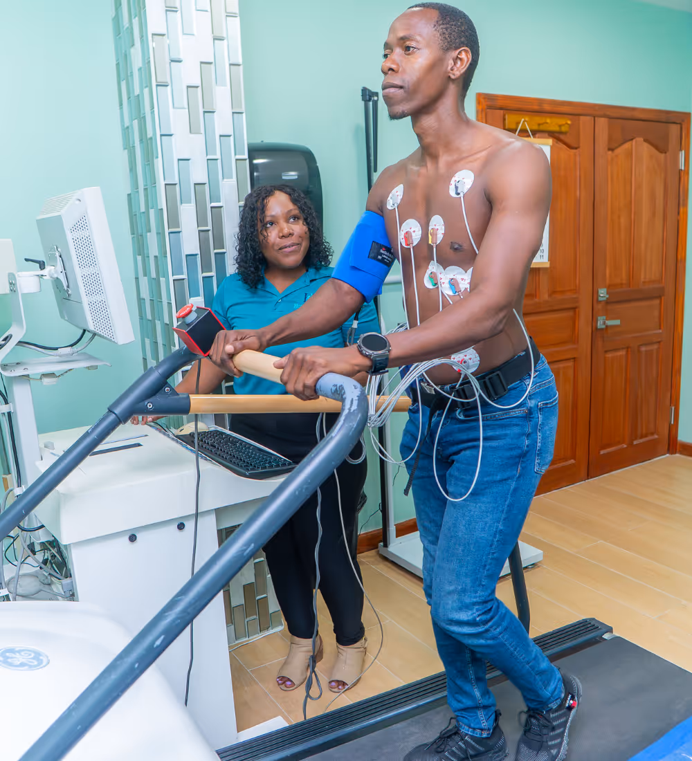 Man on treadmill with heart electrodes and blood pressure cuff, supervised by a smiling healthcare worker.