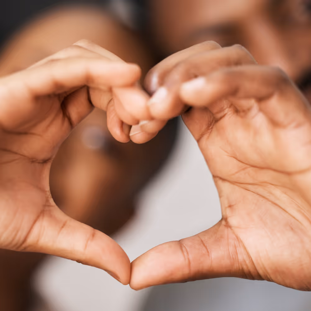 Two hands forming a heart shape with blurred person in the background.
