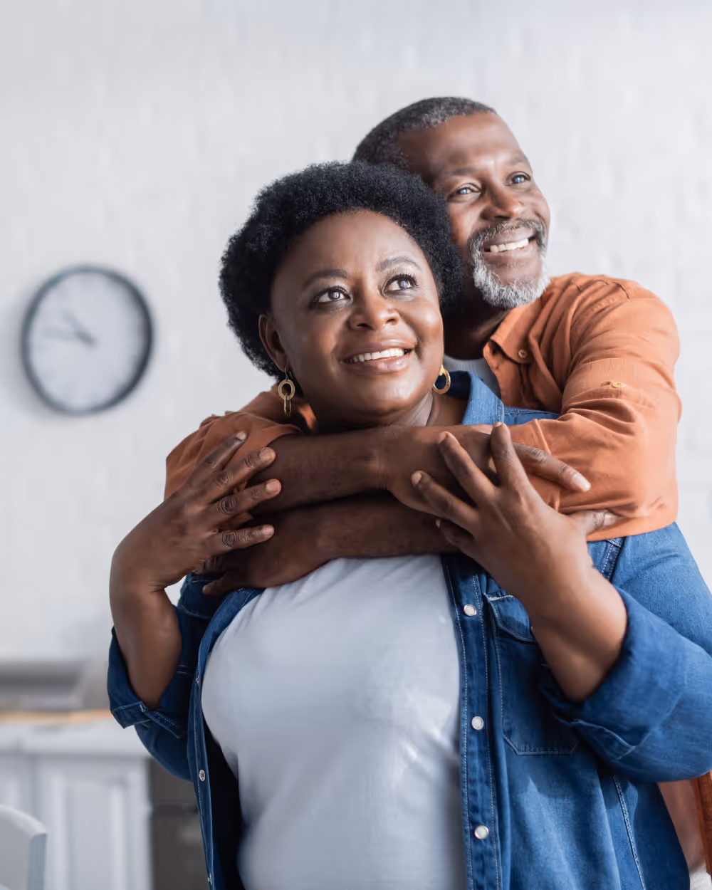 Smiling middle-aged couple hugging, with man embracing woman from behind in a home setting.