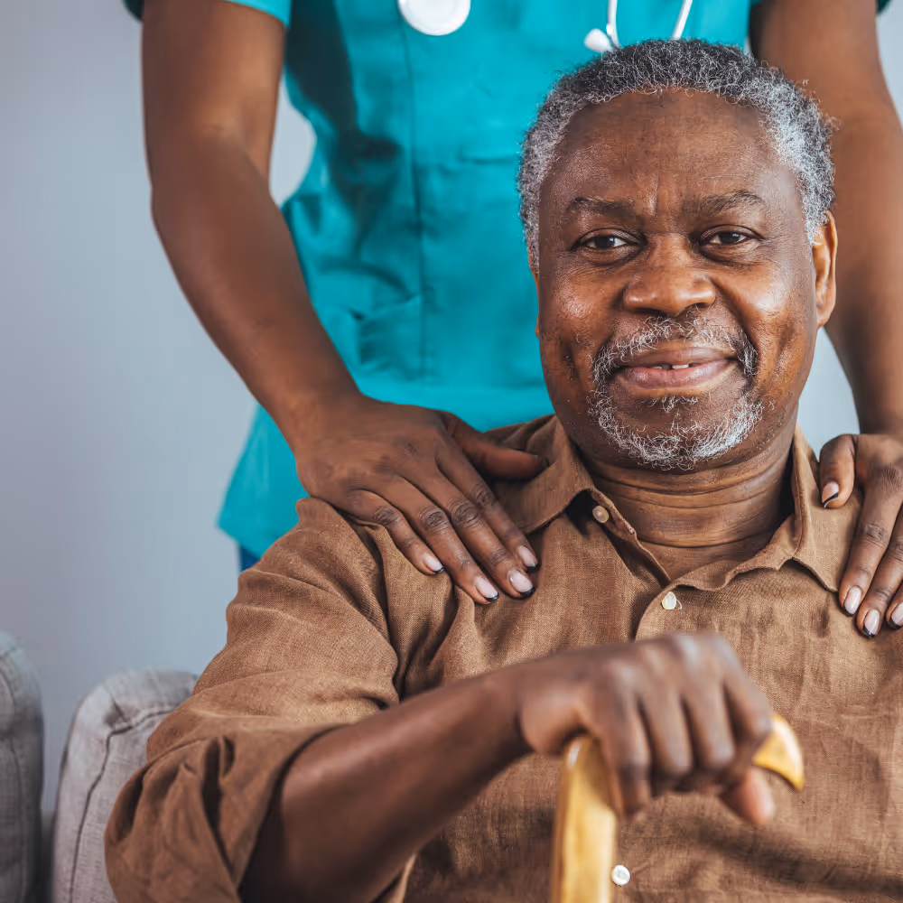 Smiling elderly man holding cane with healthcare worker in scrubs gently holding his shoulders.