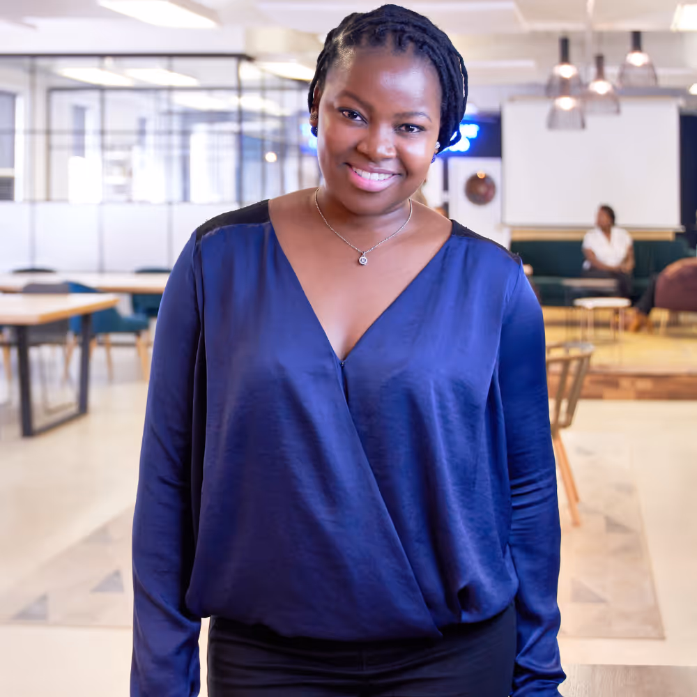 Smiling woman with braided hair wearing a blue blouse in a modern office environment.