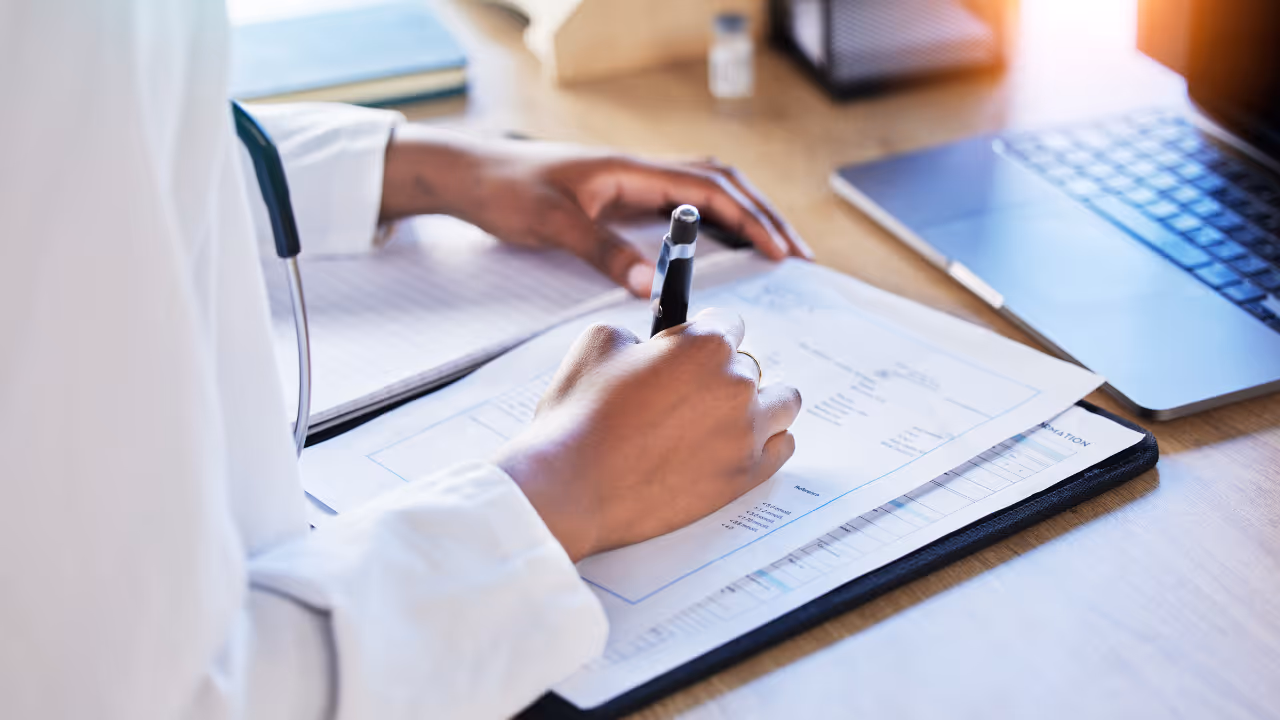 Person in white coat writing on medical document at desk with laptop and clipboard.
