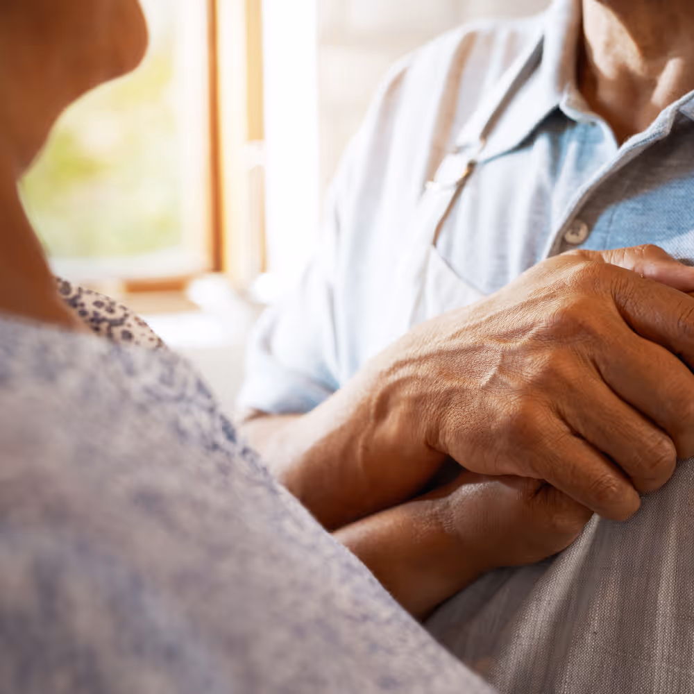 Close-up of two older adults holding hands, showing care and support indoors.