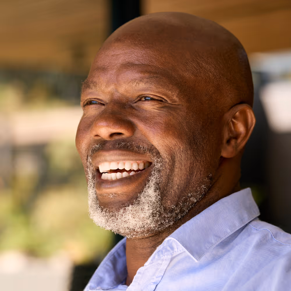 Smiling bald man with gray beard wearing a light blue collared shirt in soft natural light.