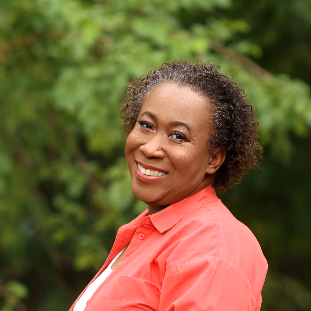 Smiling middle-aged woman with curly hair wearing a coral shirt outdoors with green foliage background.