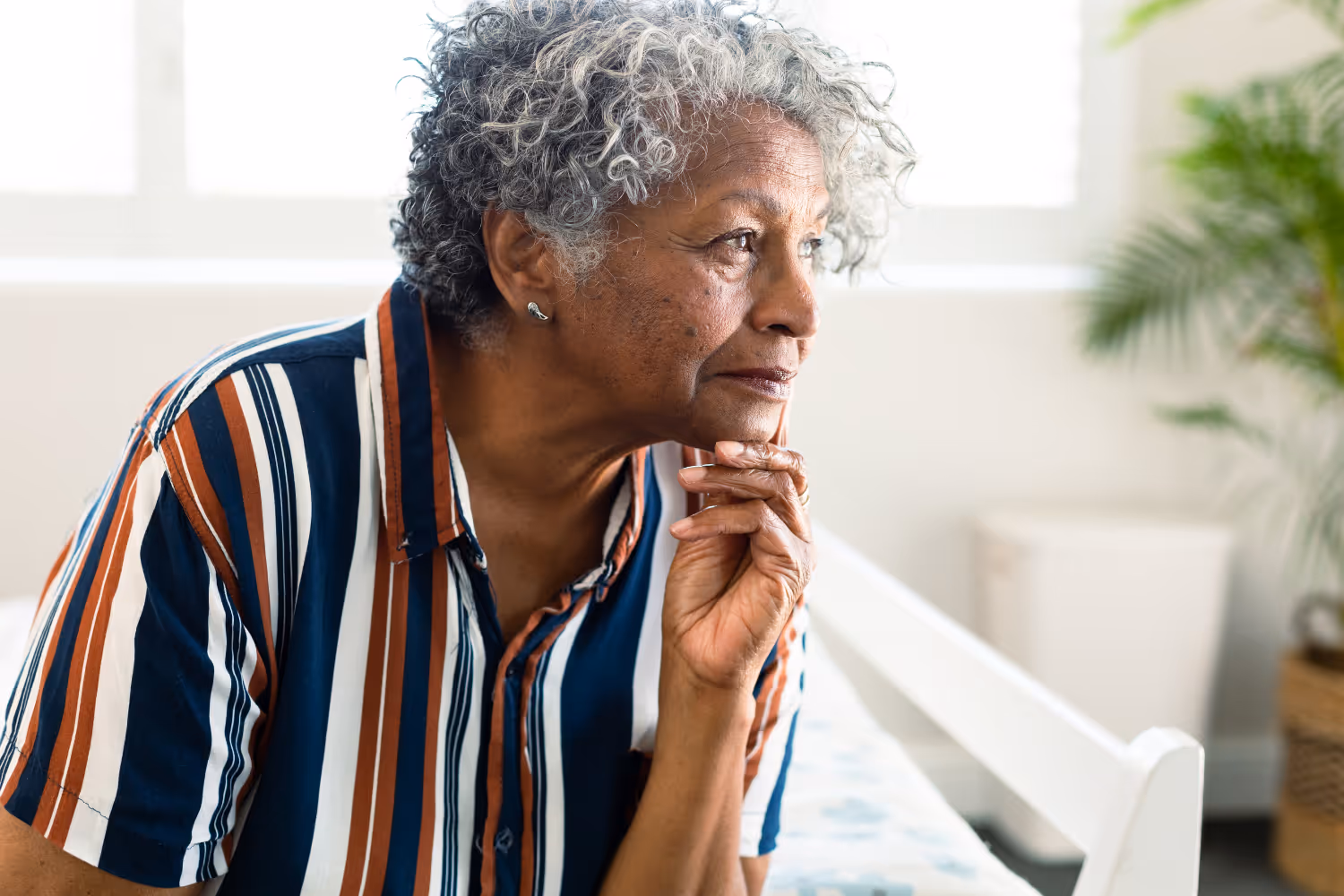 Older woman with curly gray hair in striped shirt sitting indoors, looking out with a thoughtful expression.