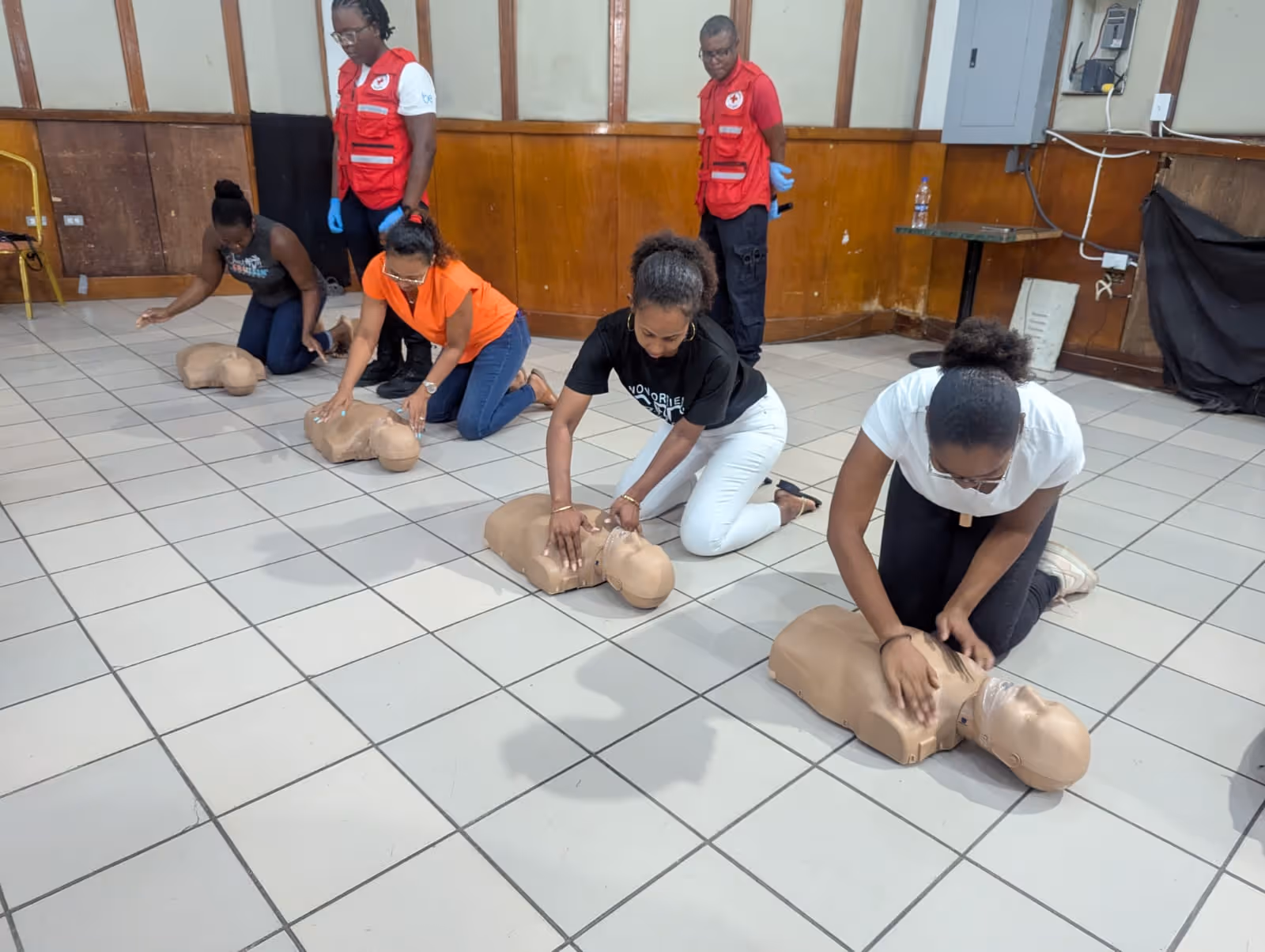 Four women kneeling on tiled floor practicing CPR on training mannequins while two instructors observe.