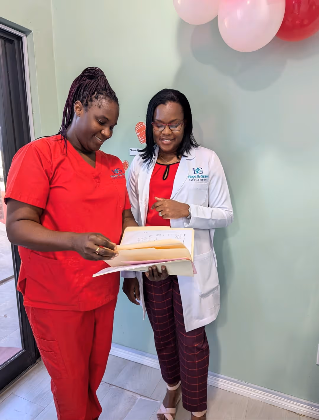 Two female healthcare workers reviewing documents, one in red scrubs, the other in white coat.