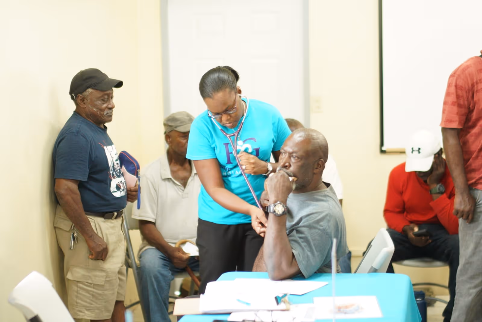 Healthcare worker uses a stethoscope to check an older man seated at a table in a clinic.