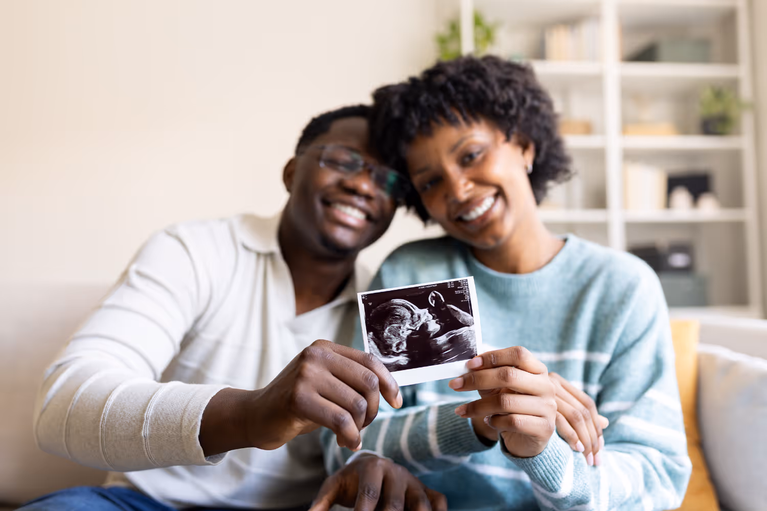 Smiling couple sitting on a couch holding an ultrasound image of their baby.