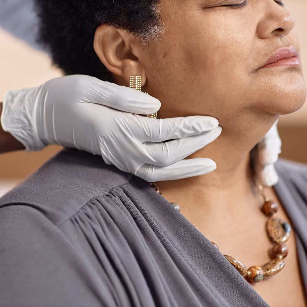 Gloved hands examining the neck of a woman wearing a gray top and beaded necklace.