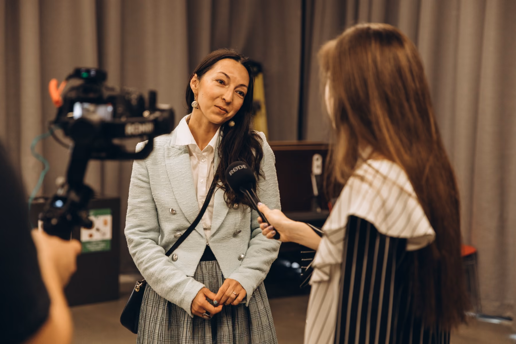 A woman in a light blazer and plaid skirt is being interviewed on camera by another Sprobuj team