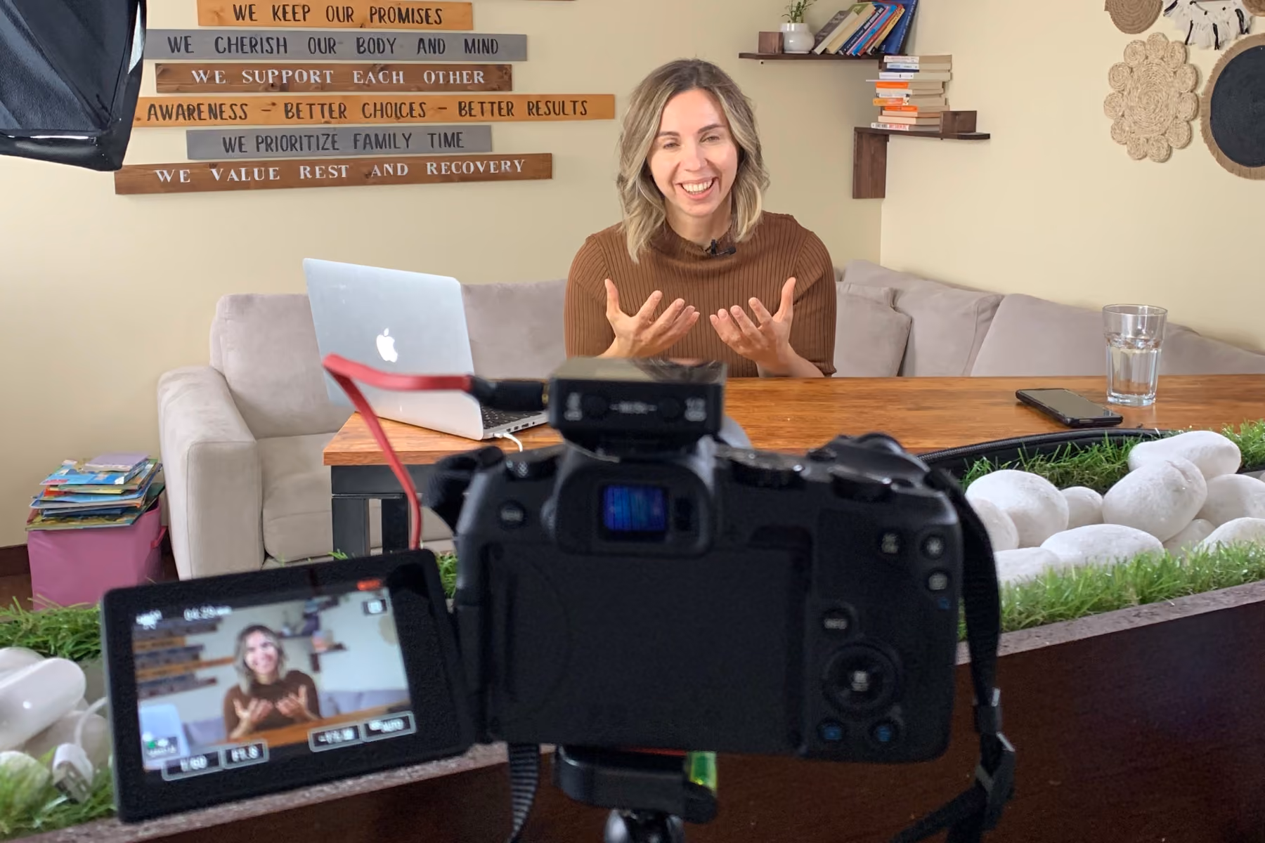 Woman with shoulder-length hair wearing a brown sweater, speaking and smiling in front of a wooden table with a laptop, recorded by a camera on a tripod in a cozy room with motivational wooden signs on the wall behind her.