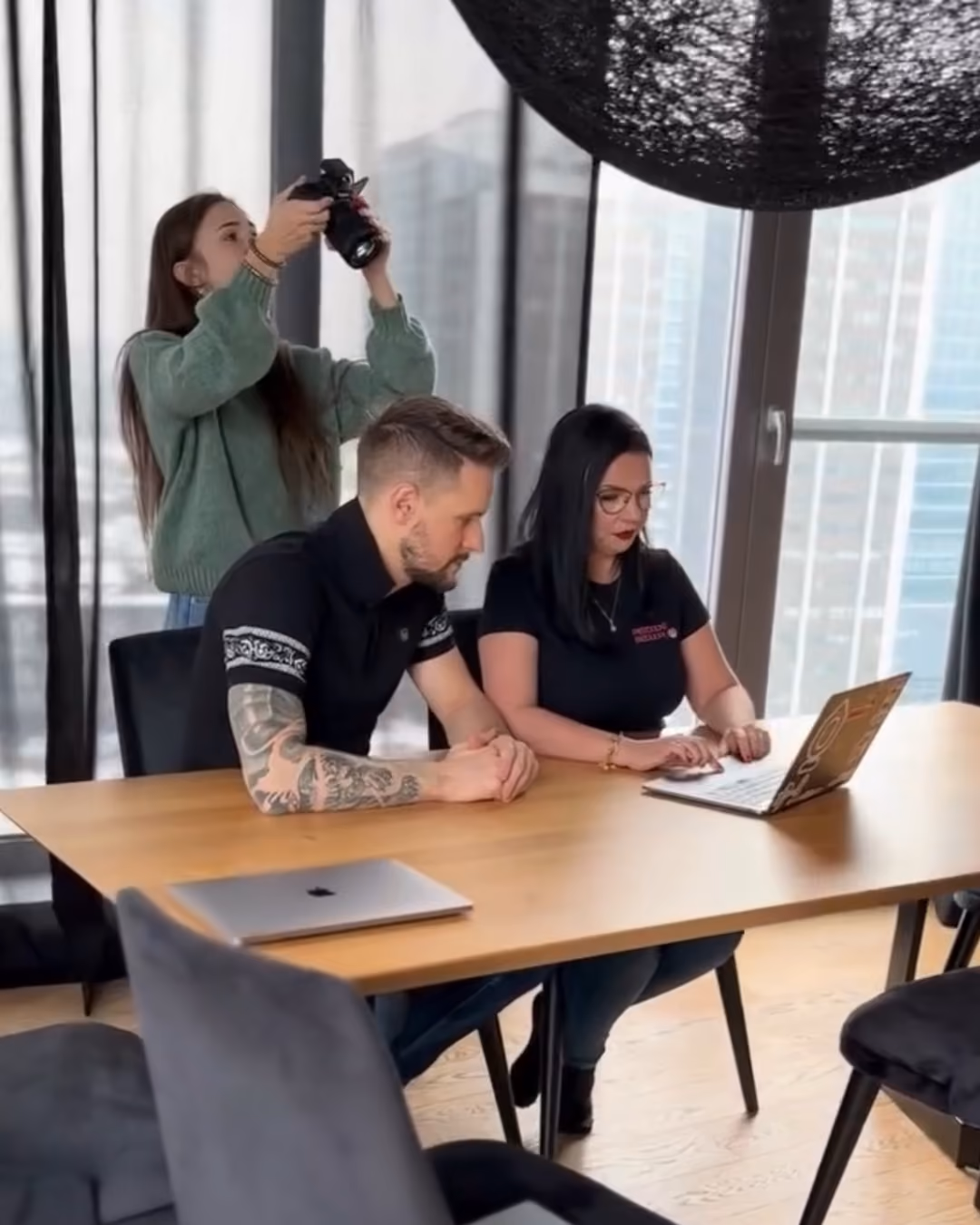 Two people sitting at a wooden table working on a laptop while a Sprobuj taking a photos