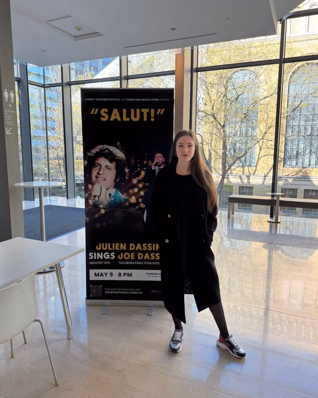 Kate stands indoors near a promotional banner for 'Salut!' featuring Julien Dassin singing Joe Dassin