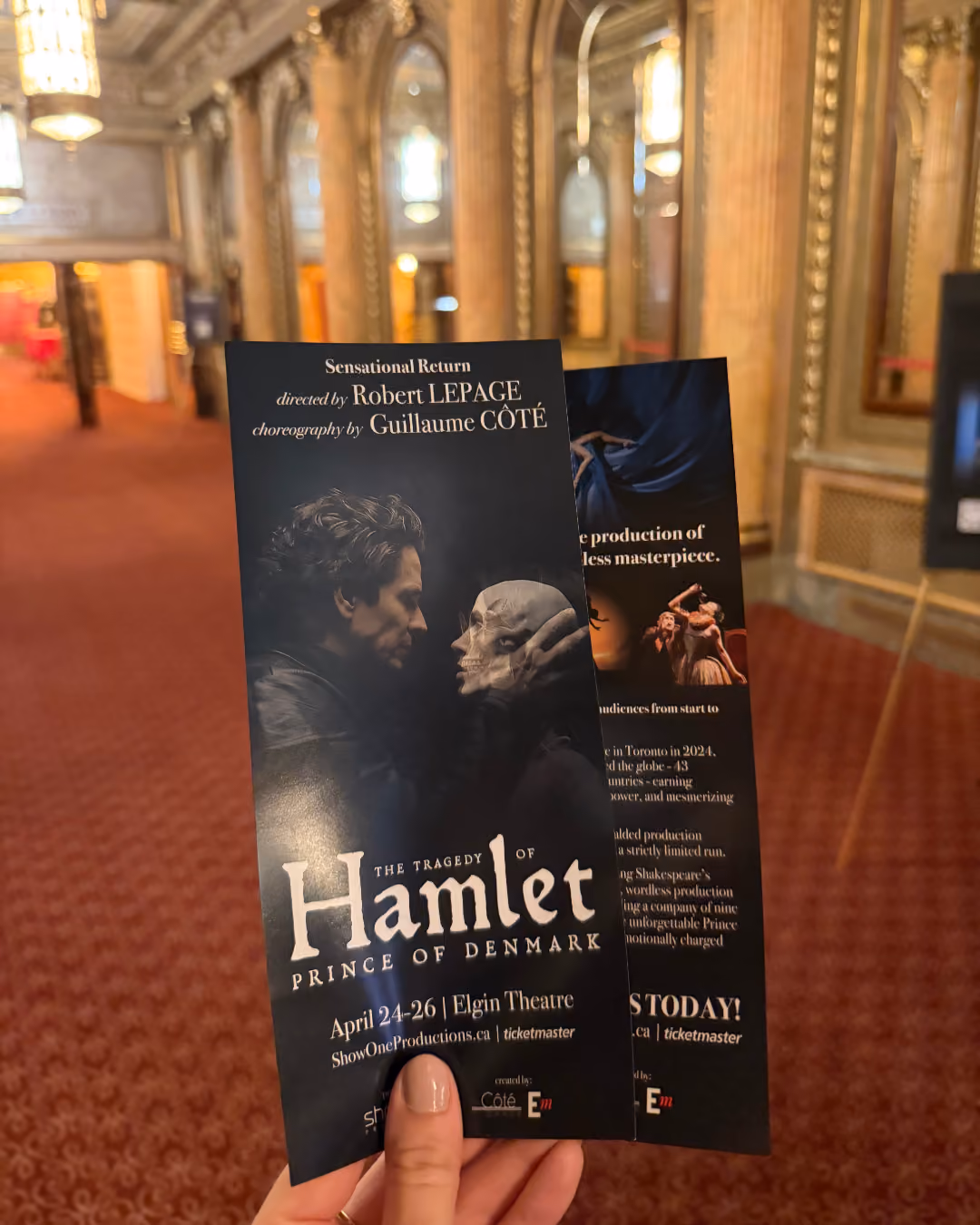 Hand holding two black flyers for the theatrical production 'The Tragedy of Hamlet, Prince of Denmark' directed by Robert Lepage and choreographed by Guillaume Côté.