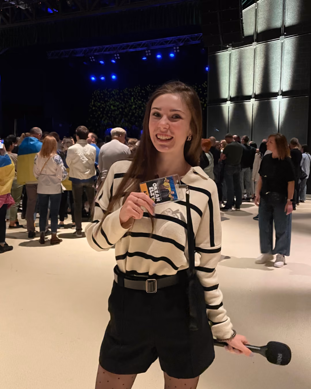 Kate holding a microphone in one hand and an event pass in the other at a crowded indoor venue with blue stage lights.